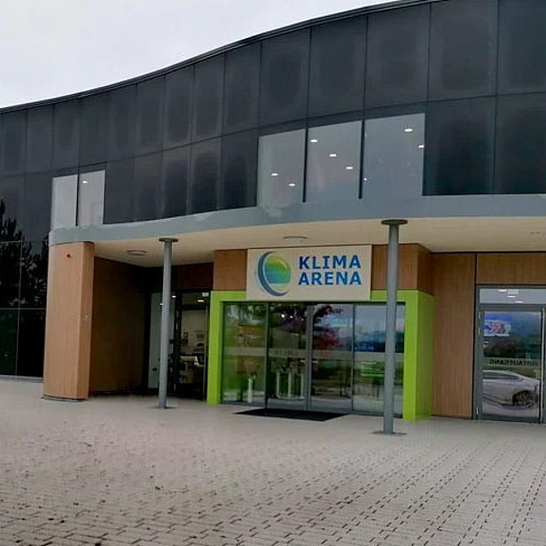 Eingang zur Klima Arena mit großer Glasfront und moderner Architektur. Links steht ein buntes, herbstliches Baum. Über der Eingangstür prangt das Logo und der Schriftzug „KLIMA ARENA“. Die Umgebung wirkt einladend und freundlich.