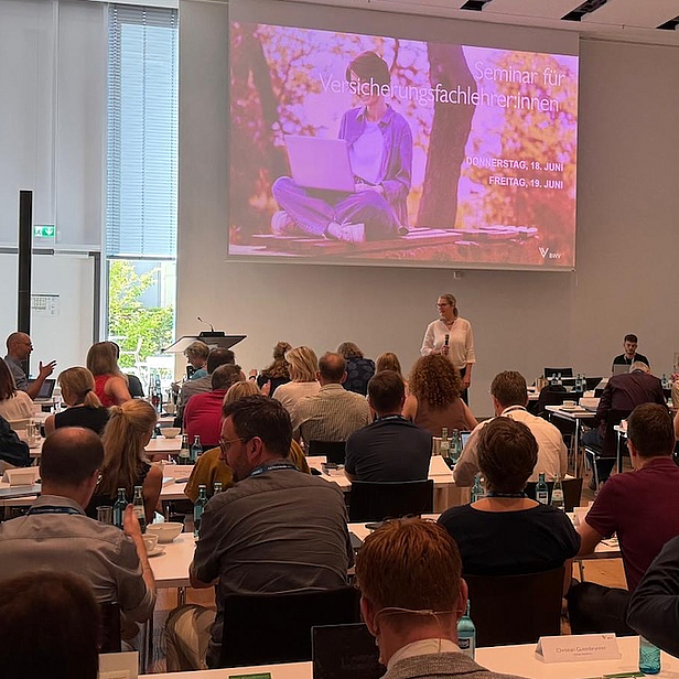 Eine Person steht vor einer Gruppe von Menschen in einem Konferenzraum und präsentiert Inhalte auf einer Leinwand. Die Teilnehmer sind aufmerksam, einige heben die Hände. Der Raum ist hell erleuchtet und bietet eine freundliche Atmosphäre für eine Schulung oder ein Seminar.