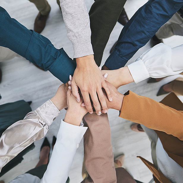 Mehrere Hände verschiedener Personen, die übereinander gestapelt sind, symbolisieren Zusammenarbeit und Teamarbeit. Die Hände zeigen unterschiedliche Hauttöne und Bekleidungsstile, was Vielfalt und Einheit darstellt. Der Fokus liegt auf dem Gemeinschaftsgefühl.