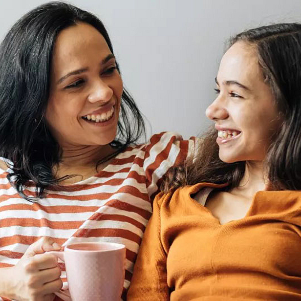 Eine Mutter und ihre Tochter sitzen auf einem Sofa und lächeln sich an. Die Mutter hält eine Tasse in der Hand, während die Tochter die Schulter ihrer Mutter umarmt. Beide strahlen Freude und Zuneigung aus. Im Hintergrund ist ein Laptop zu sehen.