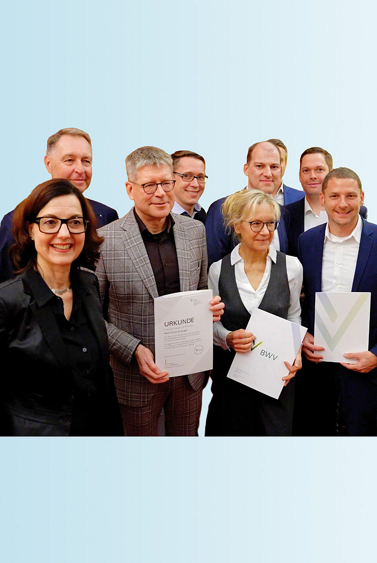 Die Abbildung zeigt eine Gruppe von acht Personen, die lächelnd zusammenstehen. Einige halten Urkunden oder Dokumente in den Händen. Im Vordergrund steht eine Frau mit Brille und dunkler Jacke. Der Hintergrund ist in sanften Blautönen gehalten.